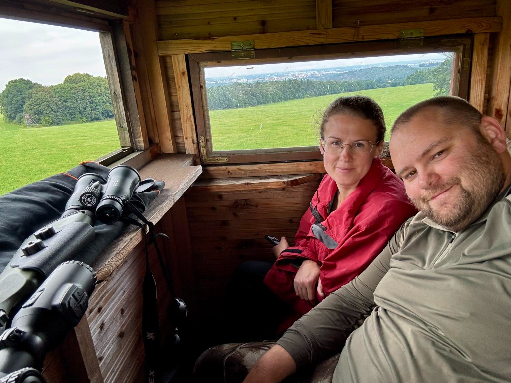 Couple smiling inside a wooden hunting blind overlooking a green field, with binoculars on the ledge