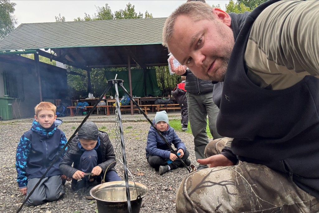 Group around a campfire with a pot on a tripod at an outdoor wooden shelter