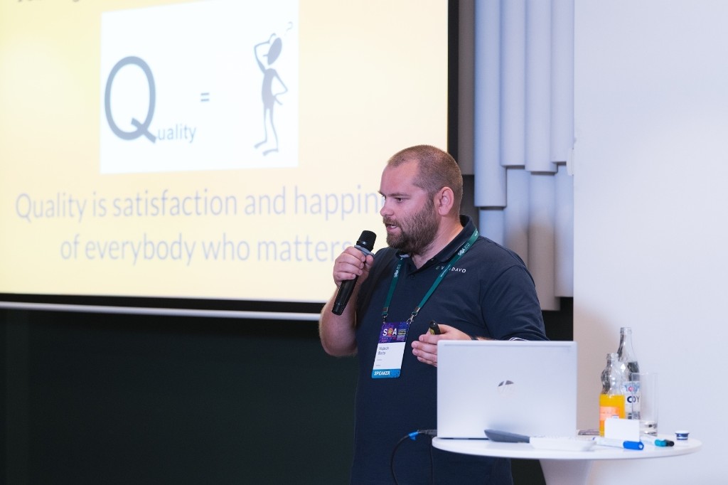 Conference talk on stage with a slide about quality and a speaker badge
